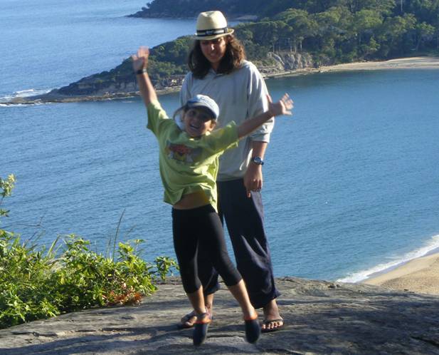 Girl bouncing up with happiness on a hilltop overlooking the beach with her big brother behind