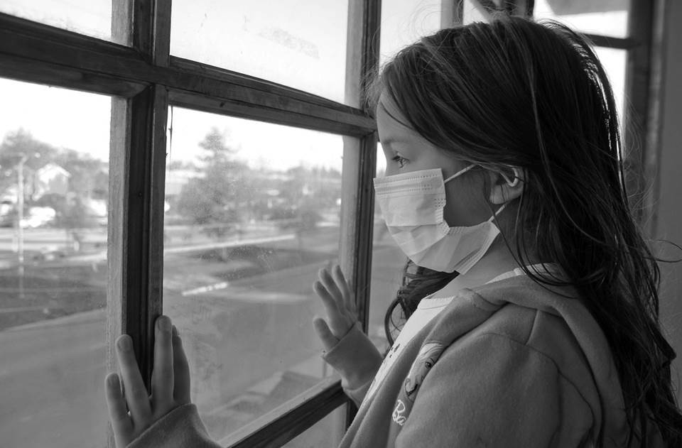 Girl in surgical mask looking sadly out the window